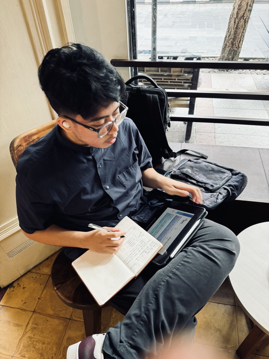 Naufal taking handwritten notes while working on his iPad at a café